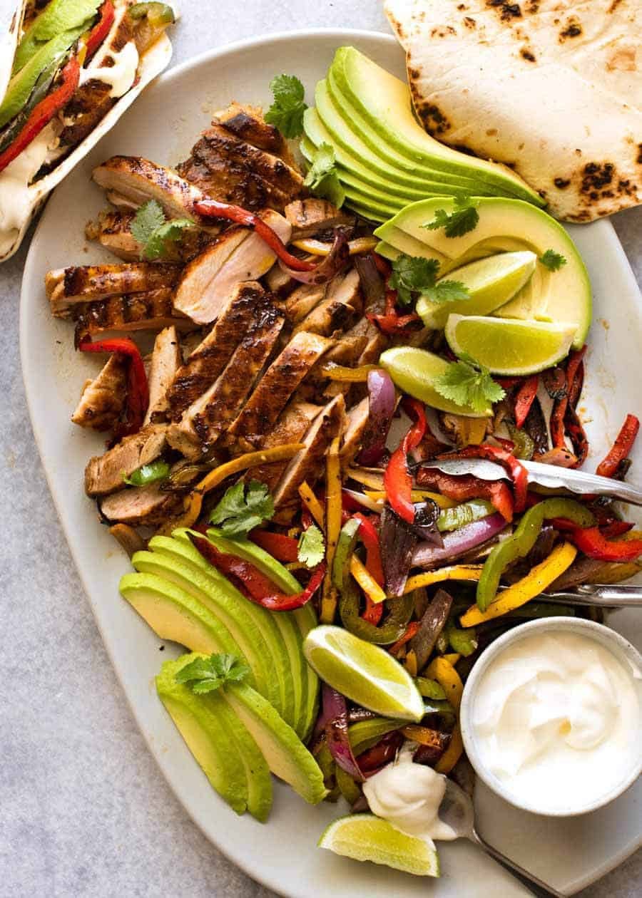 Platter of sliced Chicken Fajitas alongside charred peppers, avocado and warm tortillas, ready to be served.