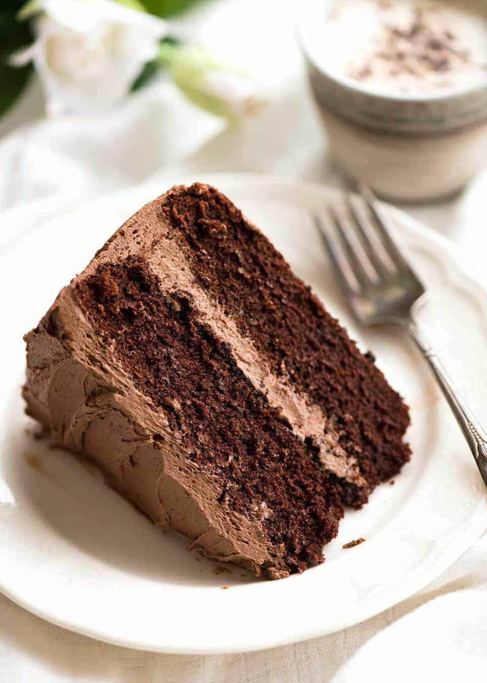 A slice of Chocolate Cake with Chocolate Buttercream Frosting on a white dessert plate with a fork, ready to be eaten.