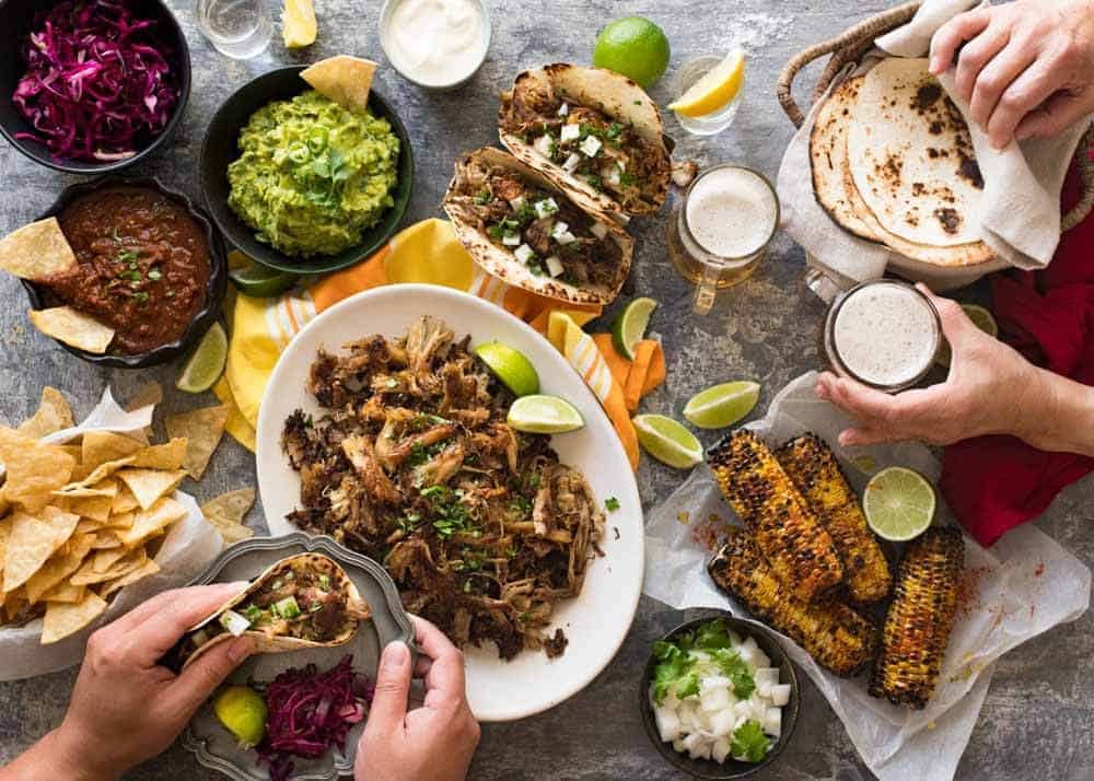 Overhead photo of Mexican Fiesta taco dinner party, with pork carnitas, tacos, grilled corn, guacamole, salsa, pickled red cabbage, tortilla chips and taco sides.